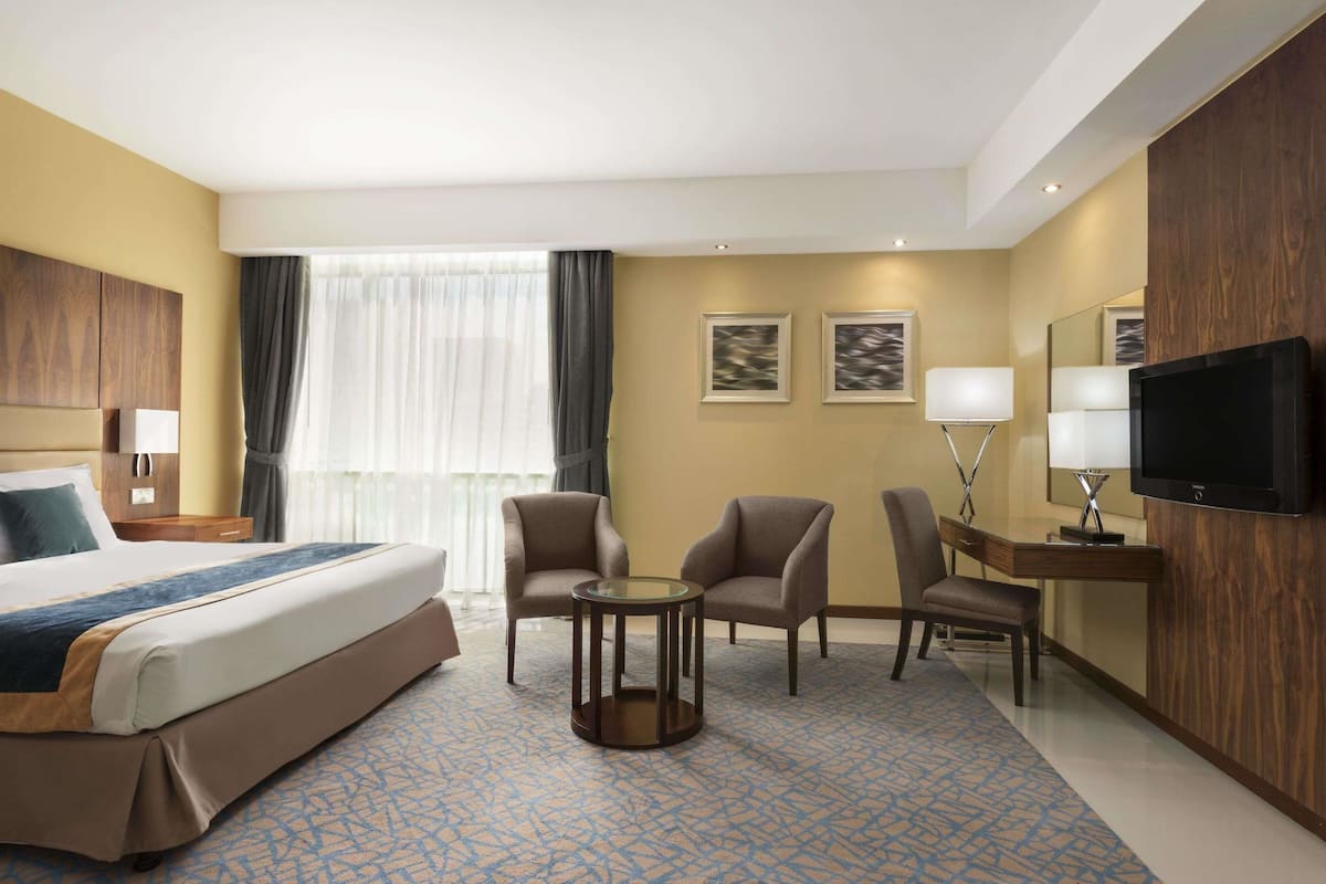Une chambre d’hôtel avec un grand lit, deux fauteuils, une petite table ronde, un bureau avec une télévision et un miroir.