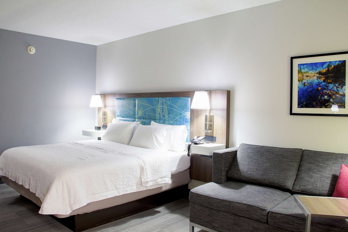 A hotel room with a bed, a grey sofa, a nightstand with a lamp, and a painting on the wall.