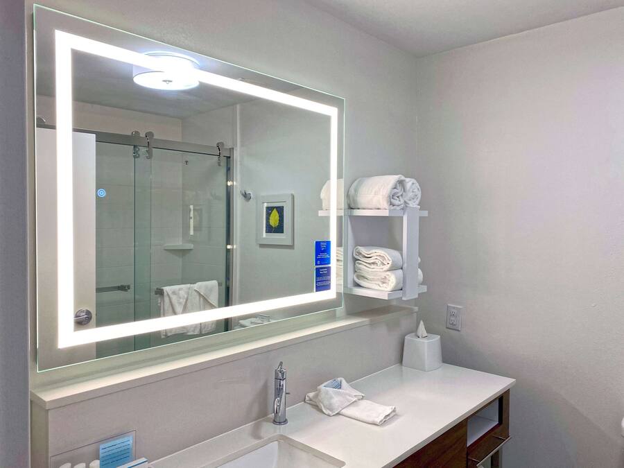 Un baño con un espejo grande, un lavamanos, un perchero para toallas y una cabina de ducha con mampara de vidrio.