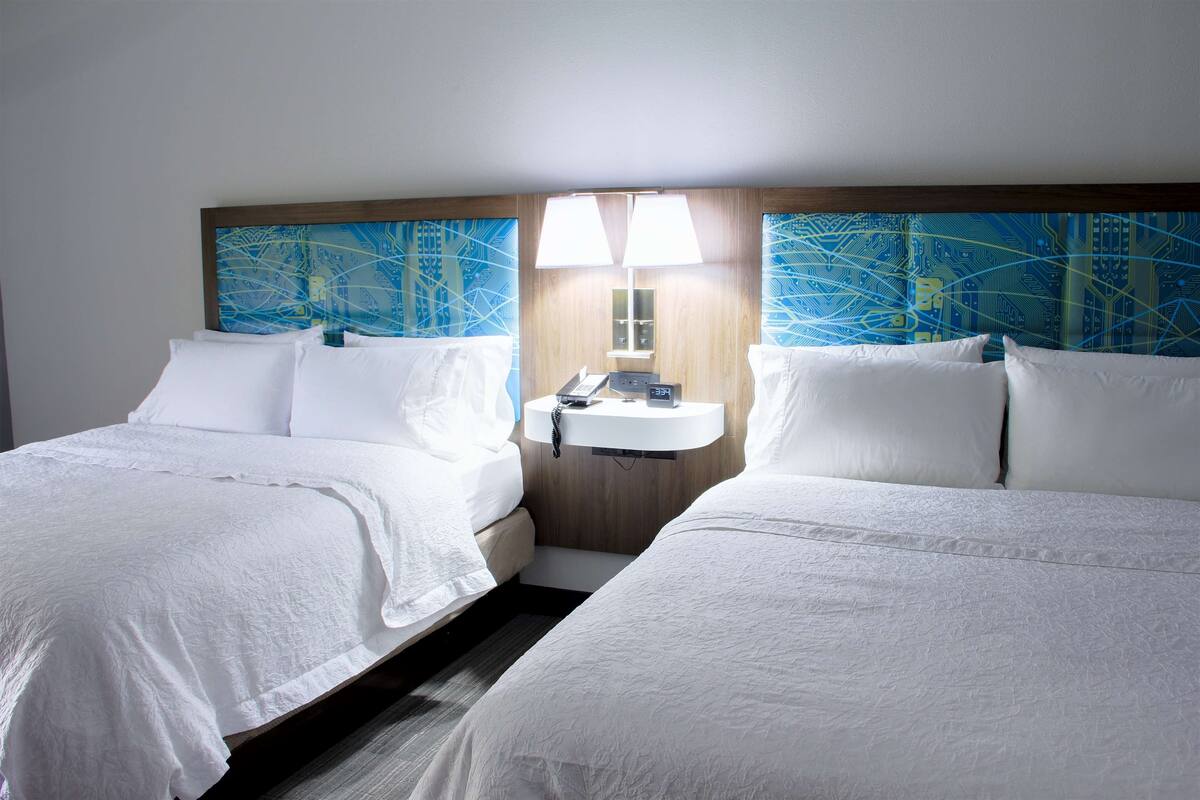 A hotel room with two beds, a nightstand with a lamp, and a wall-mounted sink.
