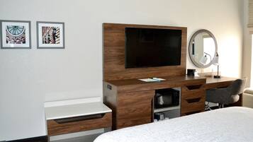 Habitación de hotel con escritorio de madera, televisor de pantalla plana, espejo, cama y dos cuadros en la pared.