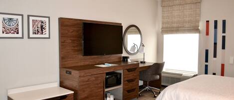 A hotel room with a wooden desk, a flat-screen TV, a mirror, a chair, and two beds.