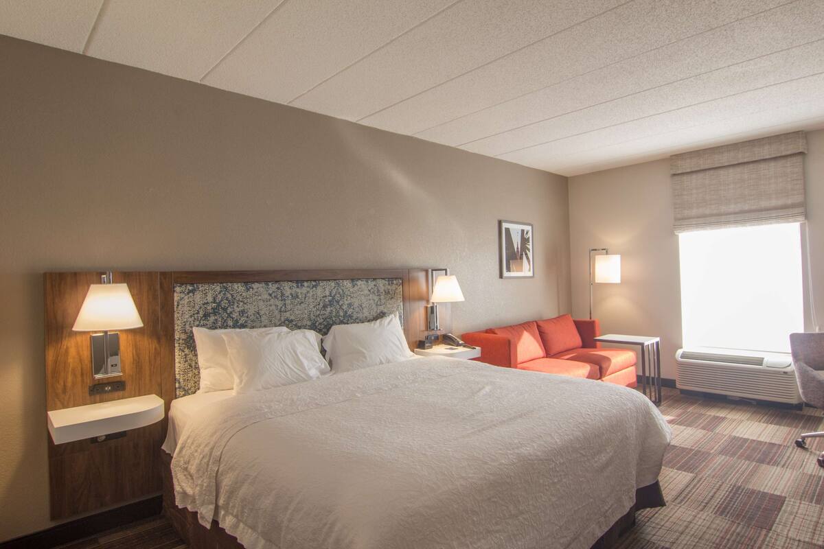 A hotel room with a large bed, two bedside lamps, a nightstand, a small table, an orange chair, and a window with a view.