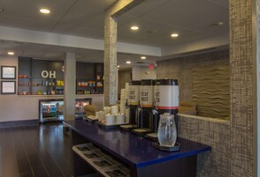 Zona de recepción de hotel con mostrador, máquinas de café, refrigerador y una pared con un cartel que dice 'OH'.