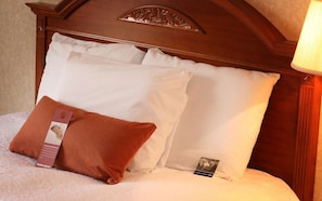 A neatly made bed with white pillows and a brown accent pillow, a wooden headboard, and a lamp on the right.