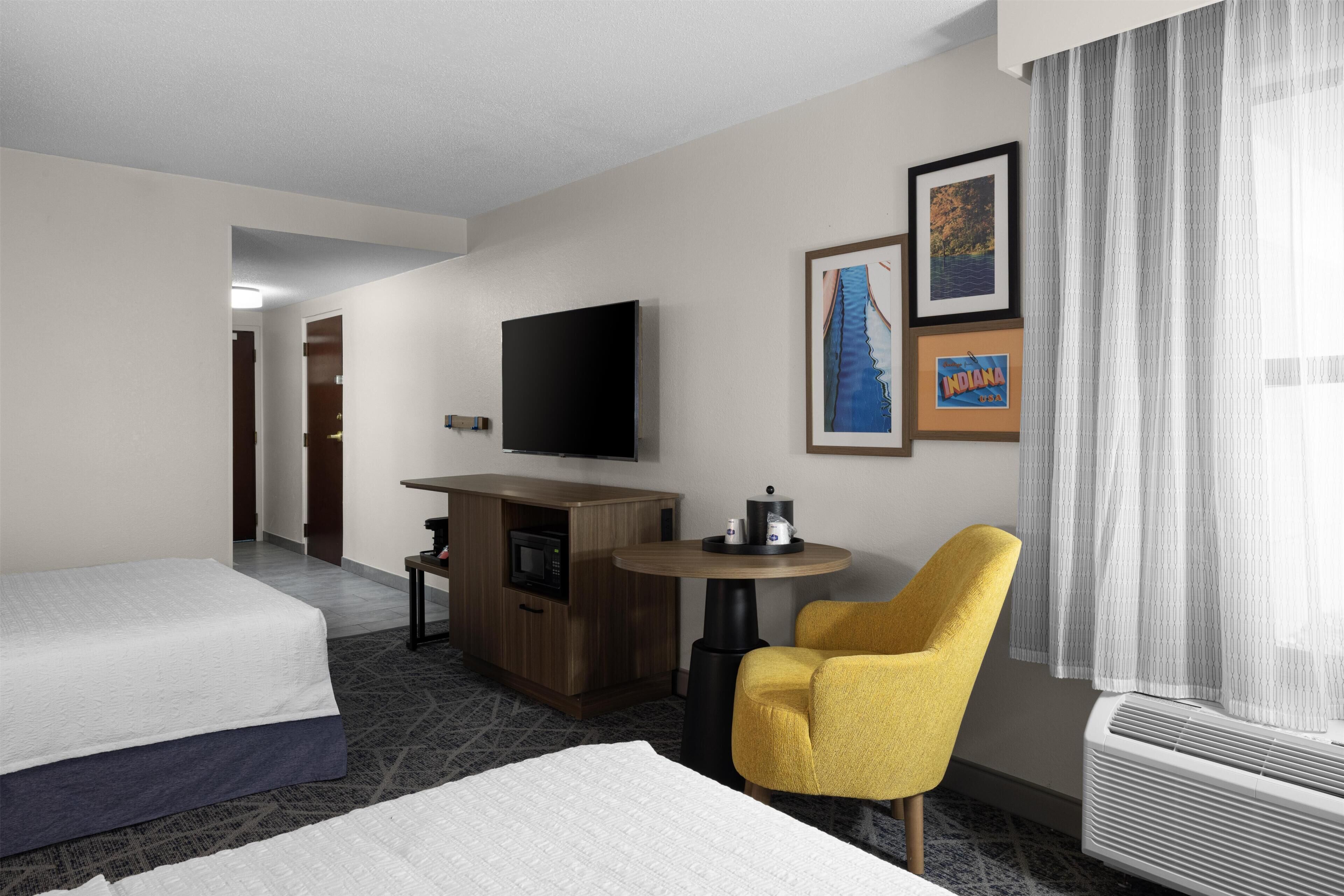 Habitación de hotel con cama, escritorio con televisor de pantalla plana, mesita, sillón amarillo y pared con cuadros.