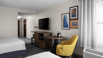 Habitación de hotel con cama, escritorio con televisor de pantalla plana, mesita, sillón amarillo y pared con cuadros.