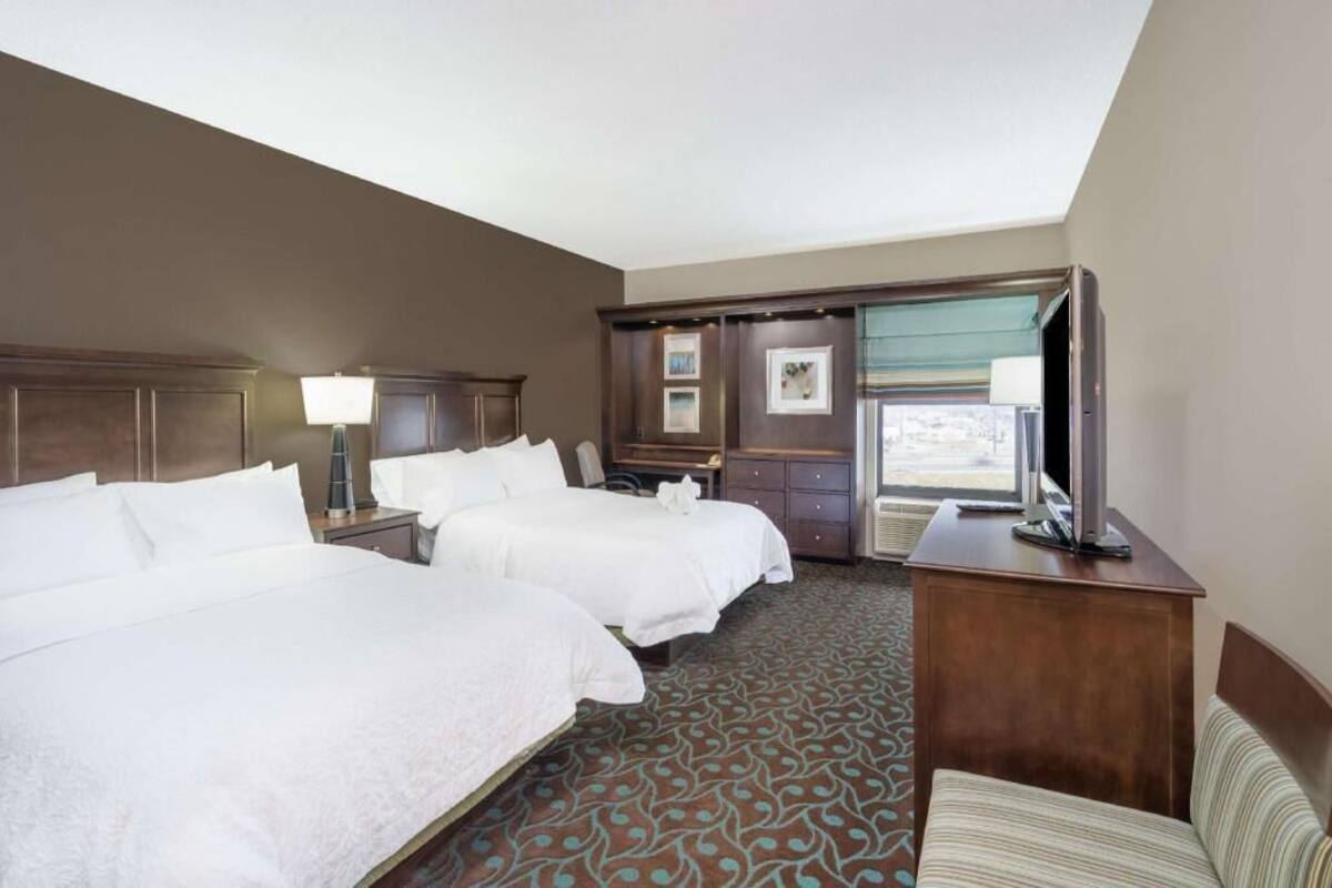 A hotel room with two beds, a TV, a desk, and a window with a view.