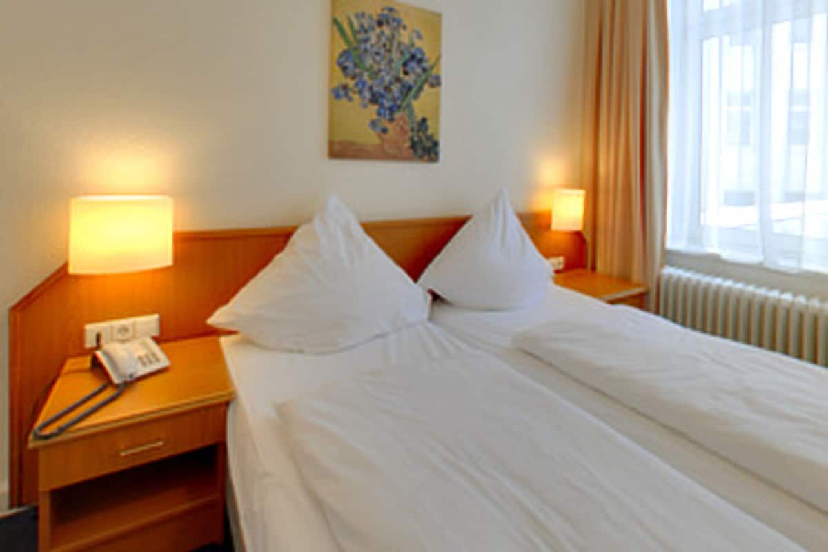 A hotel room with a neatly made bed, two bedside lamps, a nightstand with a telephone, and a window with curtains.