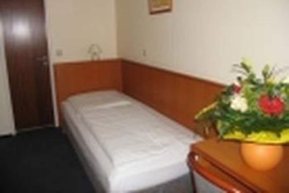 A small, single-bed room with a wooden headboard, a bedside table with a flower vase, and a door to the left.