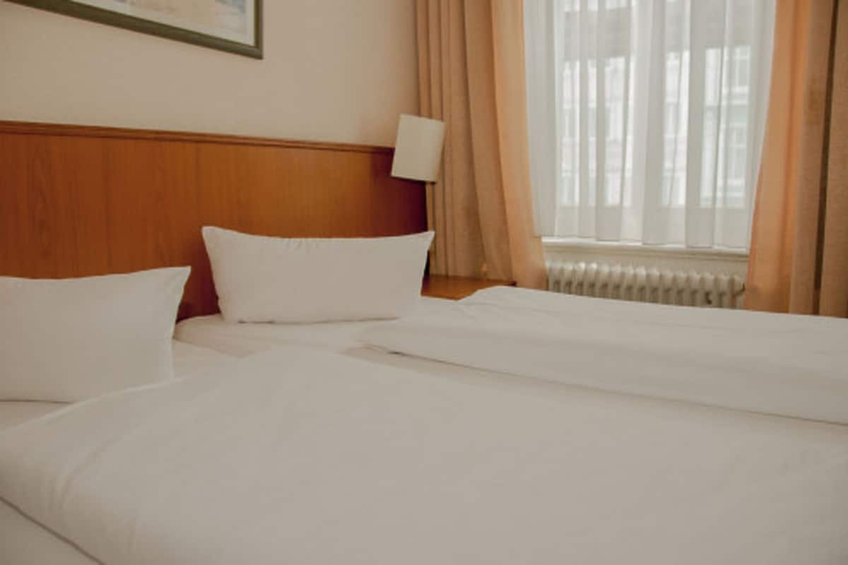 A hotel room with two beds, a wooden headboard, a wall-mounted lamp, and a window with curtains.