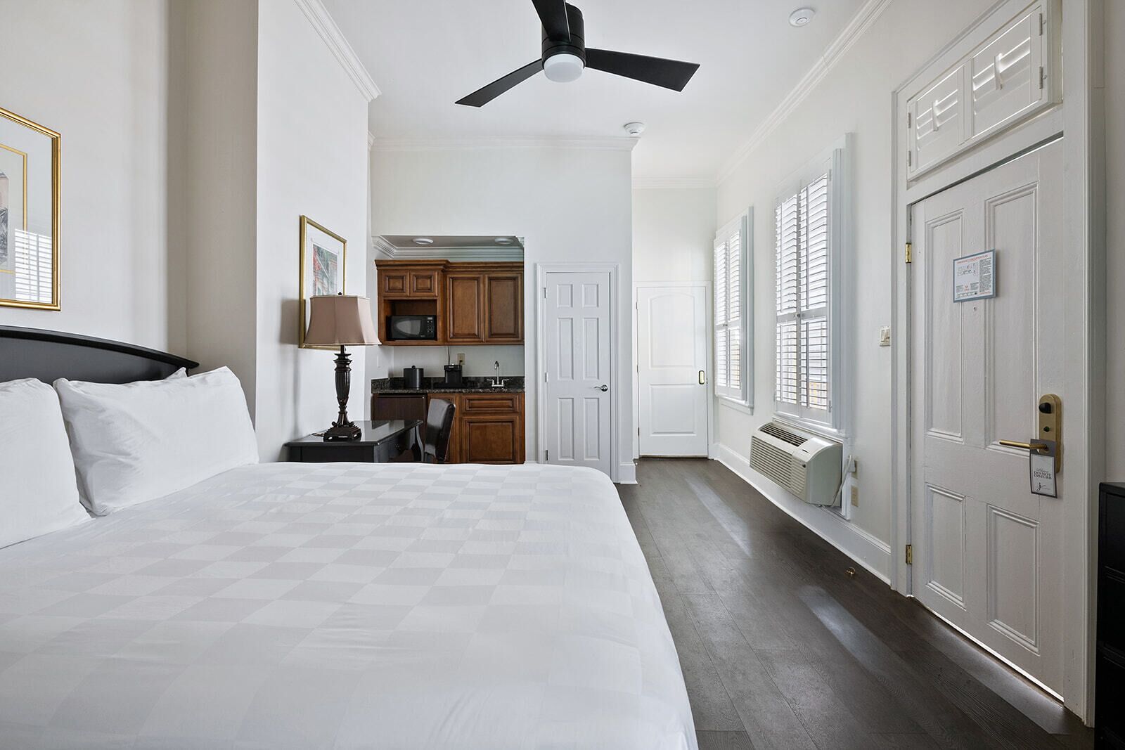 A hotel room with a large bed, a desk with a lamp, a kitchenette, and a ceiling fan.