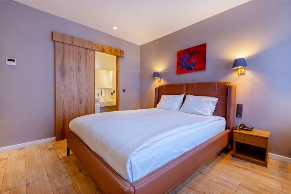 A bedroom with a large bed, wooden floor, a sliding door, a nightstand with a lamp, and a painting on the wall.