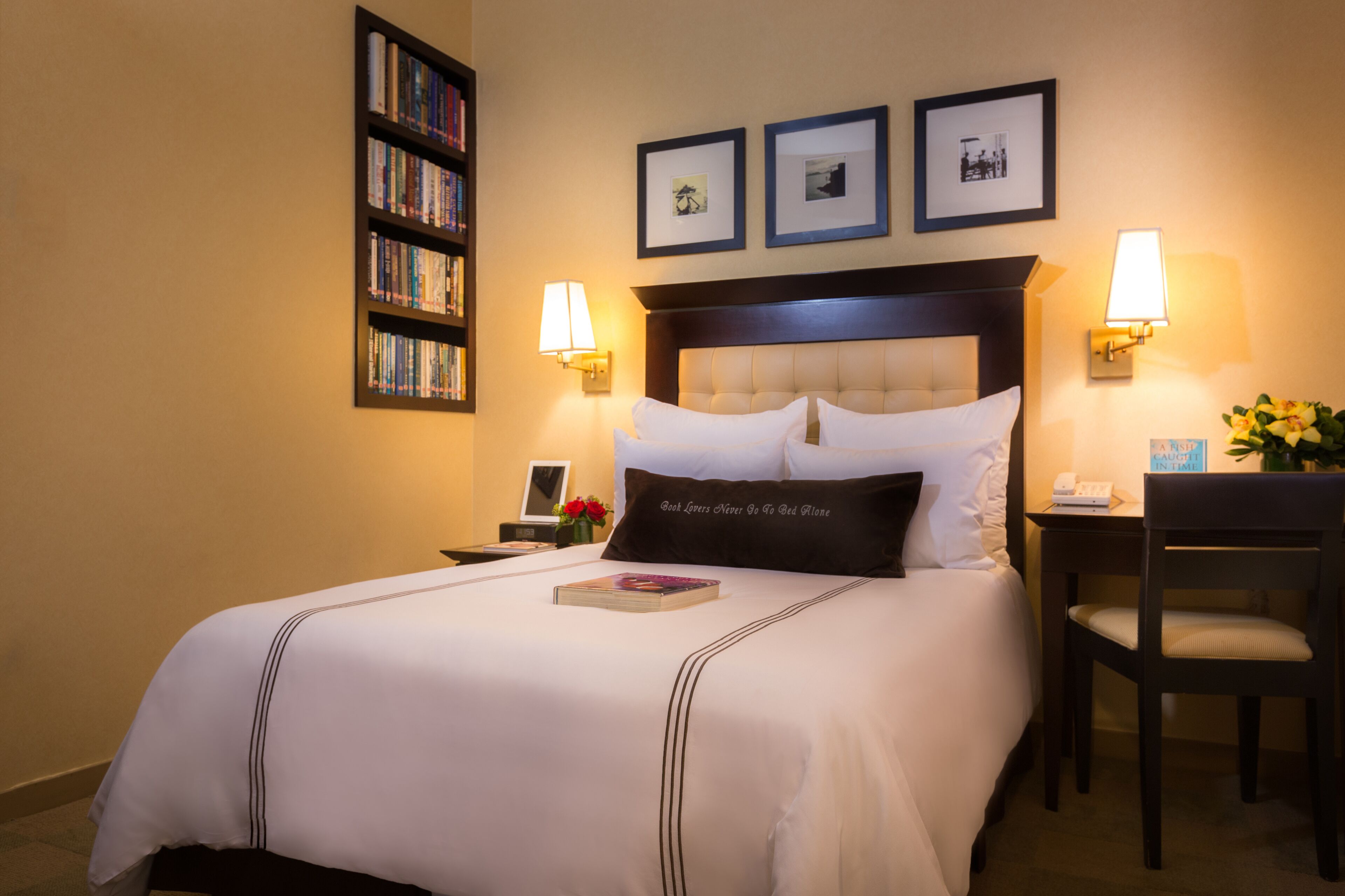 A neatly made bed with a pillow that reads "Best Lovers Never Are As Bed Alone," a bedside table with a lamp, a chair, a bookshelf, and framed pictures on the wall.
