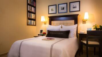 A neatly made bed with a pillow that reads "Best Lovers Never Are As Bed Alone," a bedside table with a lamp, a chair, a bookshelf, and framed pictures on the wall.