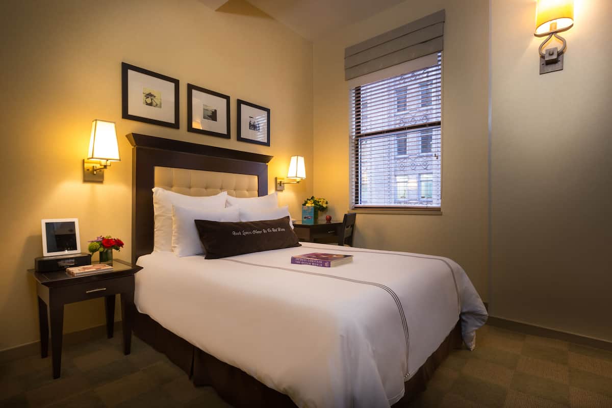 A neatly made bed with white linens, a bedside table with a lamp, a small table with a vase of flowers, and framed pictures on the wall.