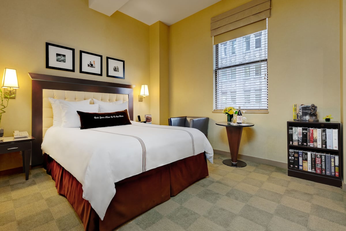 A hotel room with a large bed, two bedside lamps, a small round table with a vase of flowers, a bookshelf with books, and a window with blinds.