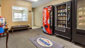 Um saguão de hotel com uma máquina de venda automática de Coca-Cola, um banco e um tapete de entrada com o nome do hotel.