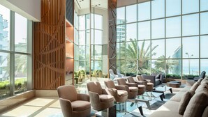 A spacious lobby with a modern design, featuring a seating area with sofas and armchairs, a glass coffee table, and large windows offering a view of palm trees and a building.