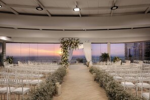 An indoor venue with rows of white chairs arranged for an event, a floral archway, and a view of the sunset through large windows.