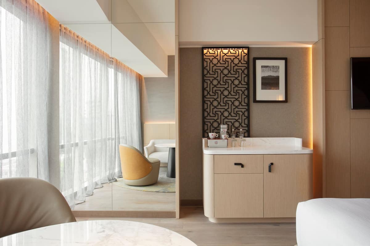 A modern hotel room with a large window, a round table, a sofa, a kitchen area with a sink, and a decorative wall panel.