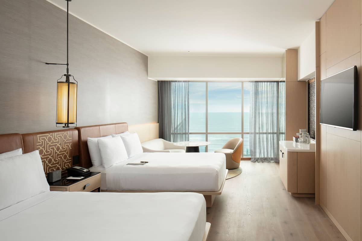 A hotel room with two beds, a TV, a desk, and a view of the sea.