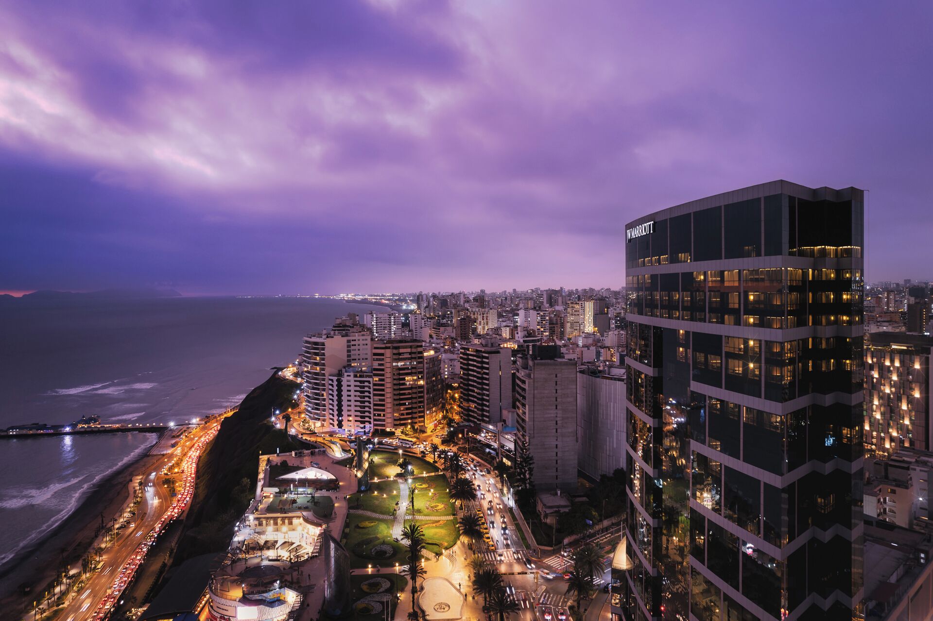 A cityscape at dusk with a prominent hotel named 'MARriott' featuring a modern design, a beachfront location, and a busy street with traffic.