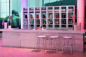 Una zona de bar con un estante blanco que muestra diversas botellas y vasos, tres taburetes de bar y una pared decorativa.