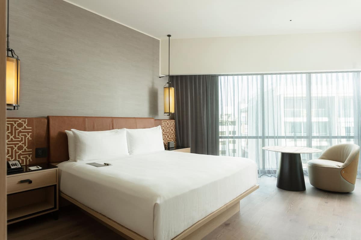 A hotel room with a large bed, bedside tables, a chair, and a view of the city through large windows.