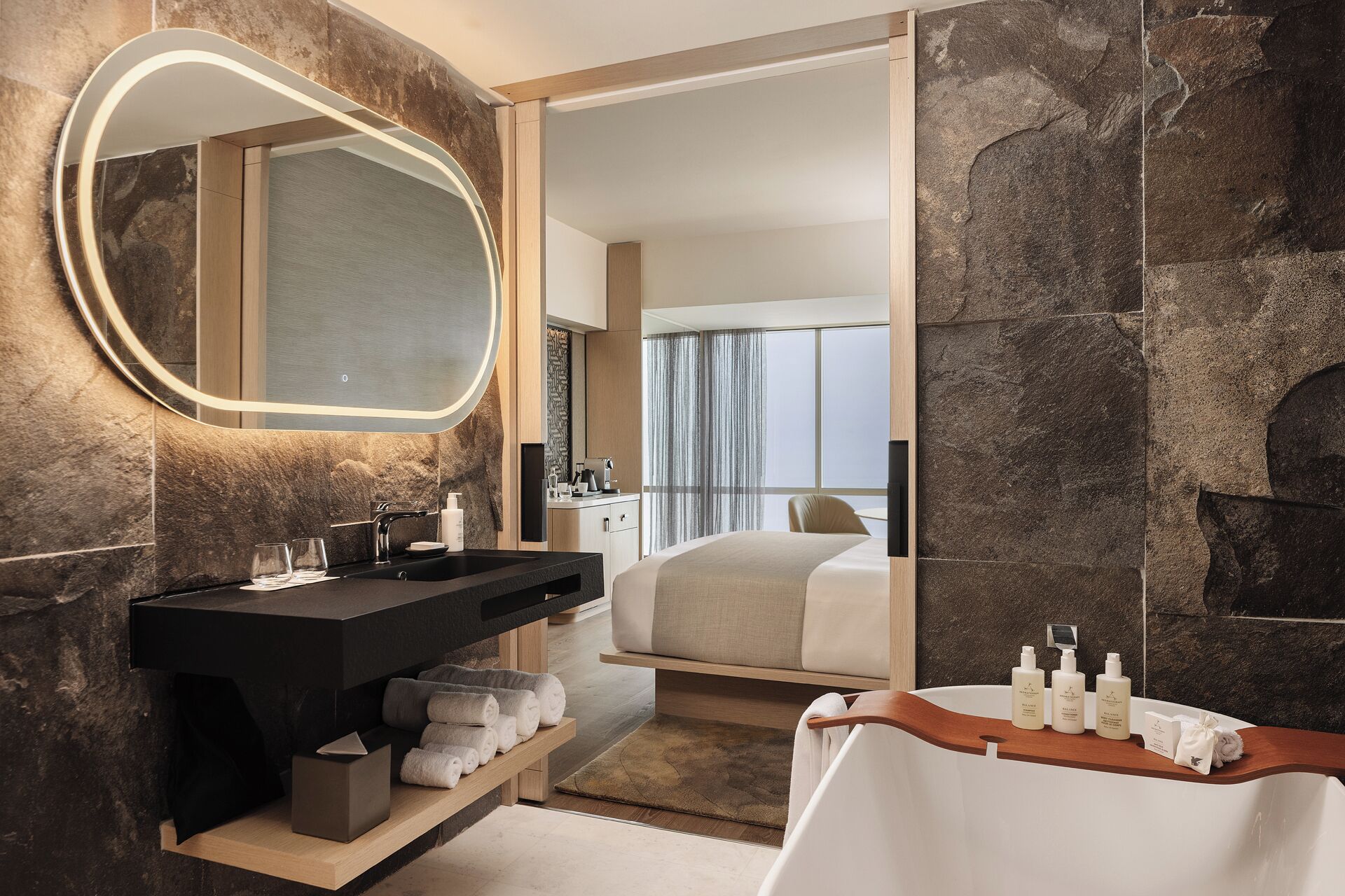 Una habitación de hotel moderna con pared de piedra, un espejo grande, tocador negro, cama y una mesita de noche de madera con artículos de aseo.