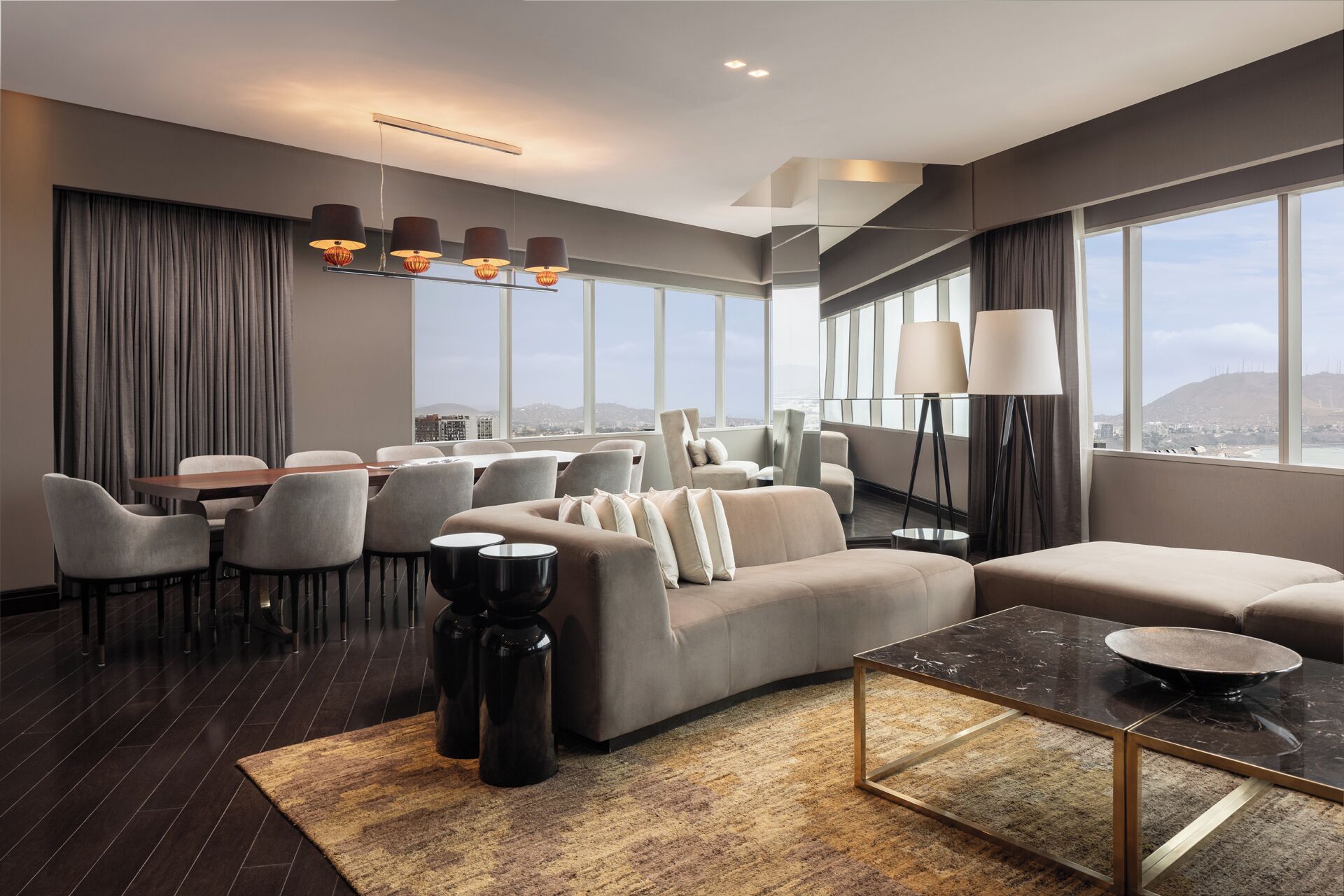 A modern hotel room with a sofa, dining area, and large windows offering a city view.