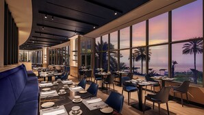 A restaurant with tables set for dining, large windows offering a view of palm trees and the sunset.