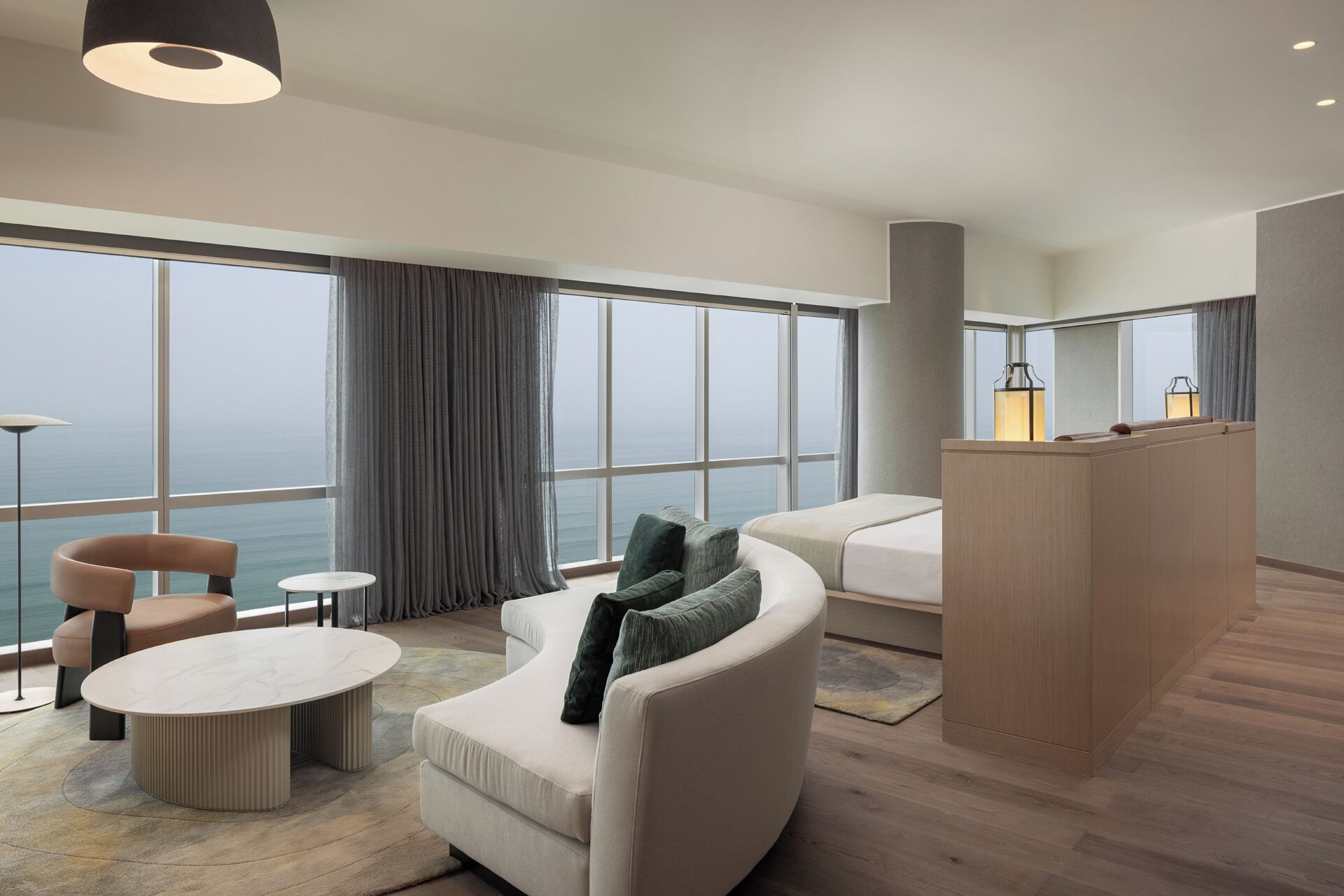 A modern hotel room with a curved sofa, a round coffee table, a small side table, a wooden desk, and large windows offering a view of the sea.