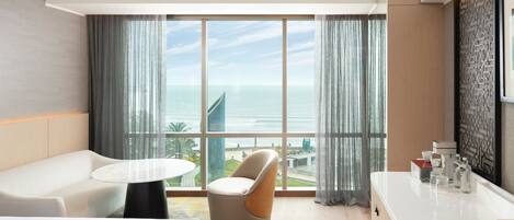 A modern hotel room with a large window offering a view of the sea and a cityscape. There is a white sofa, a round table, and a unique chair.