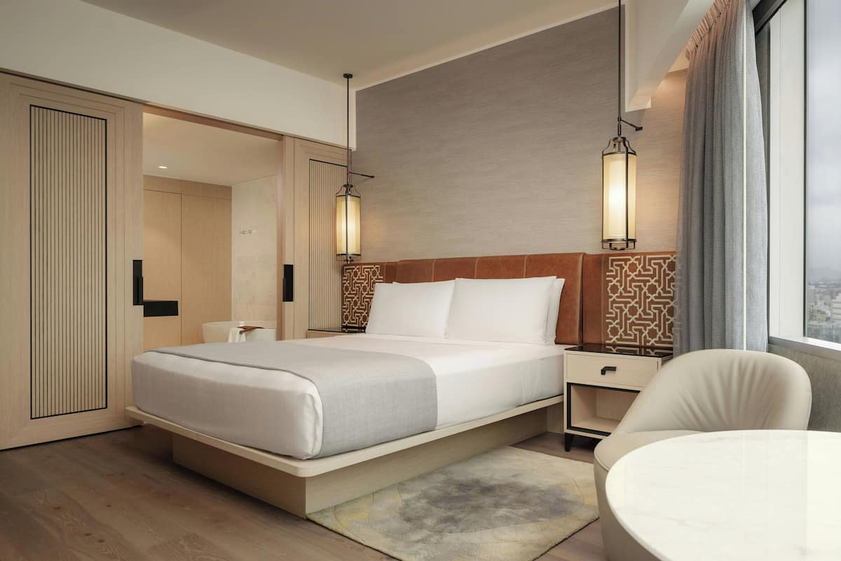 A modern hotel room with a bed, bedside table, chair, and a view of the city.
