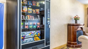 Máquina expendedora con refrigerios y bebidas, ubicada en una habitación con un escritorio de madera y una silla.
