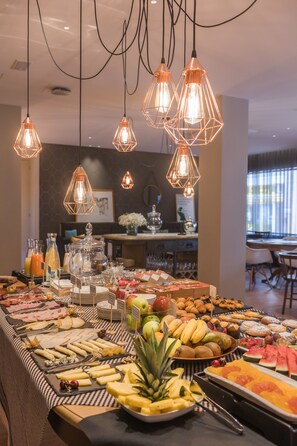 A buffet table with a variety of fruits, cheeses, and pastries, and a bar area in the background. - Up Rooms Vic Hotel (Vic)