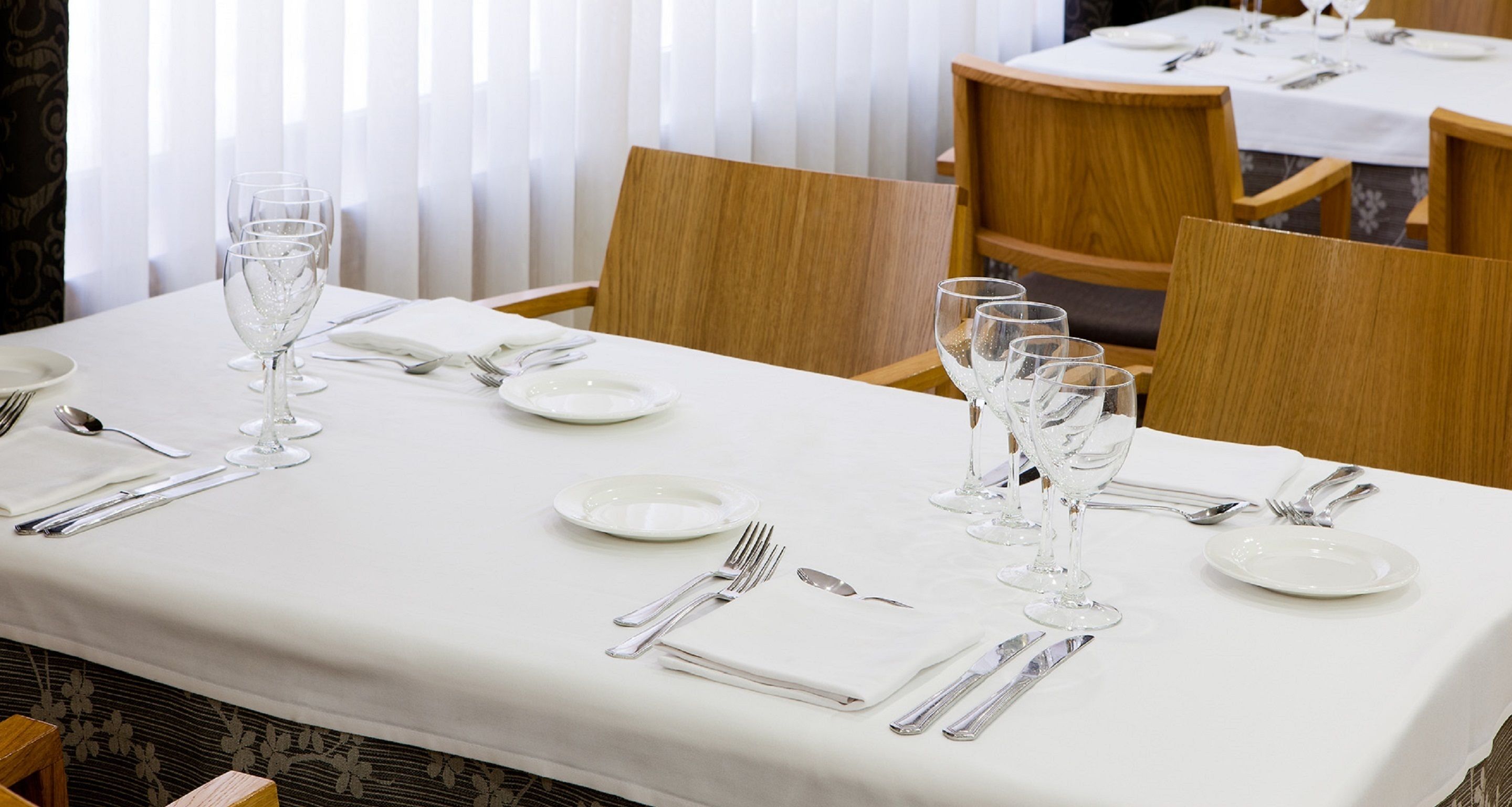 Une table à manger dressée avec une nappe blanche, des couverts en argent et des verres à vin, entourée de chaises.