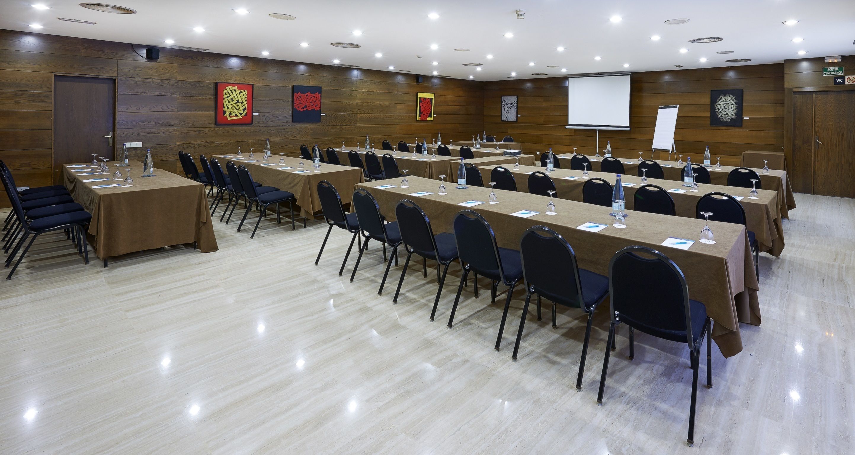 A conference room with multiple tables and chairs, equipped with water bottles and glasses, and a projector screen.