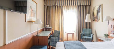 Habitación de hotel con cama, escritorio, silla, banco, televisor y cortinas.