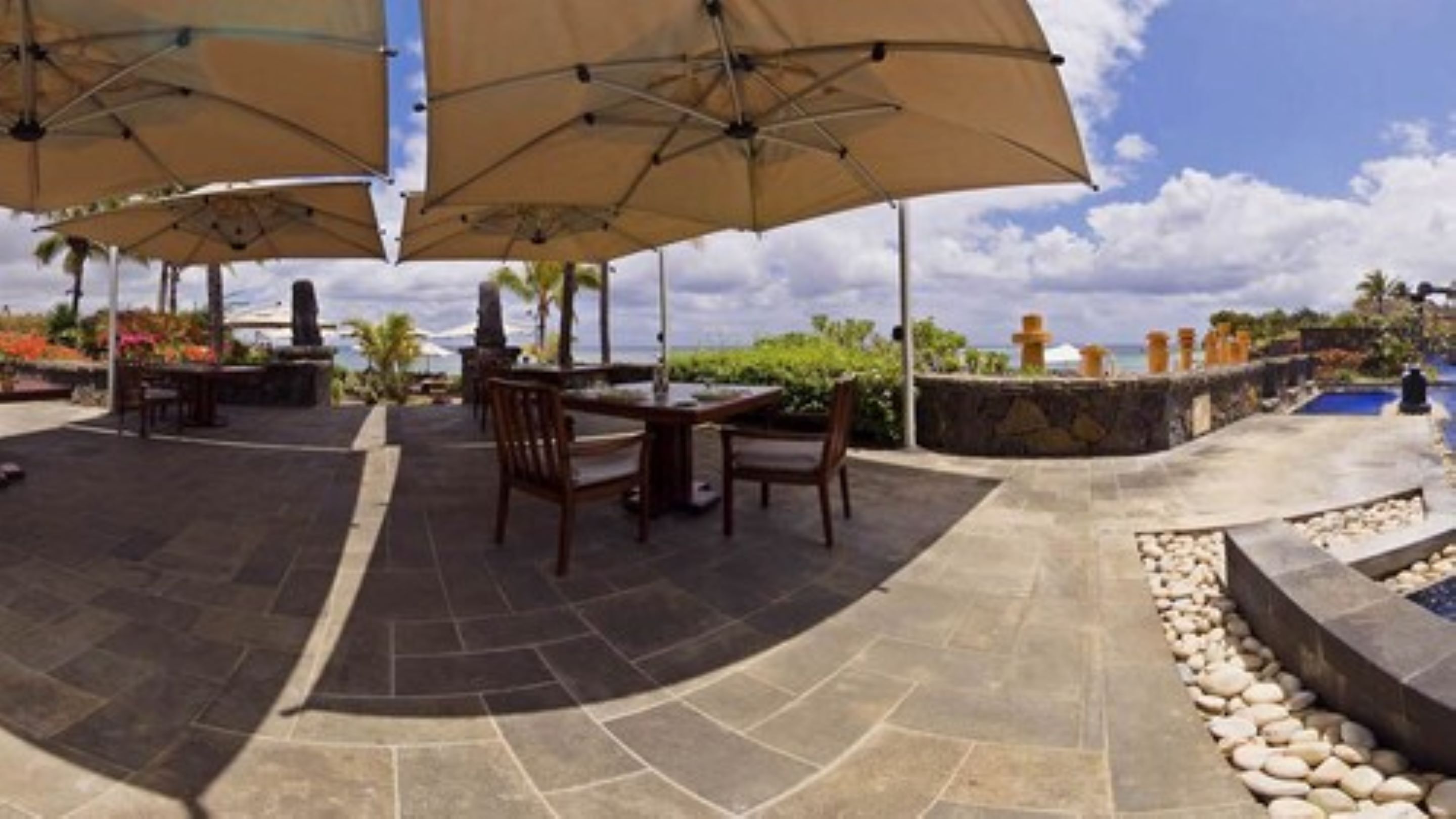 An outdoor patio area with stone flooring, wooden tables and chairs, and large beige umbrellas providing shade.