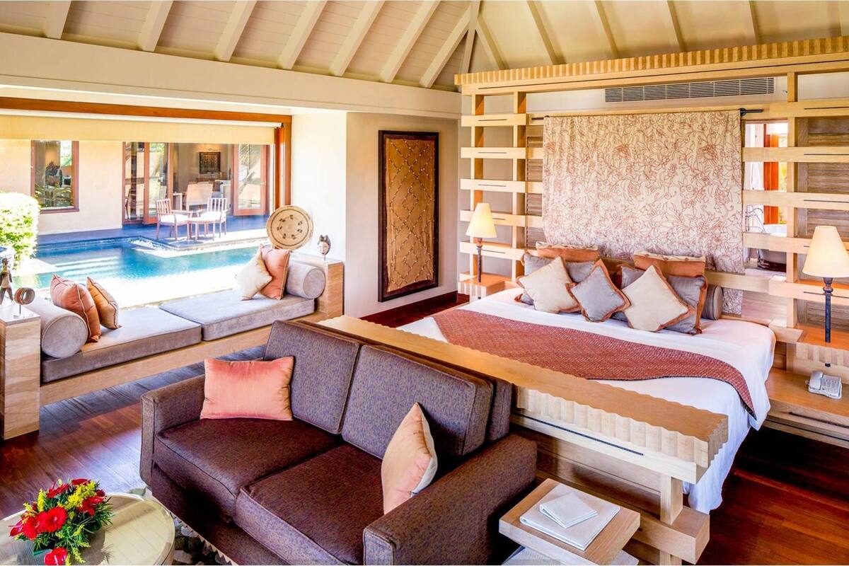 A bedroom with a bed, a sofa, a small table, a lamp, and a view of an outdoor swimming pool.