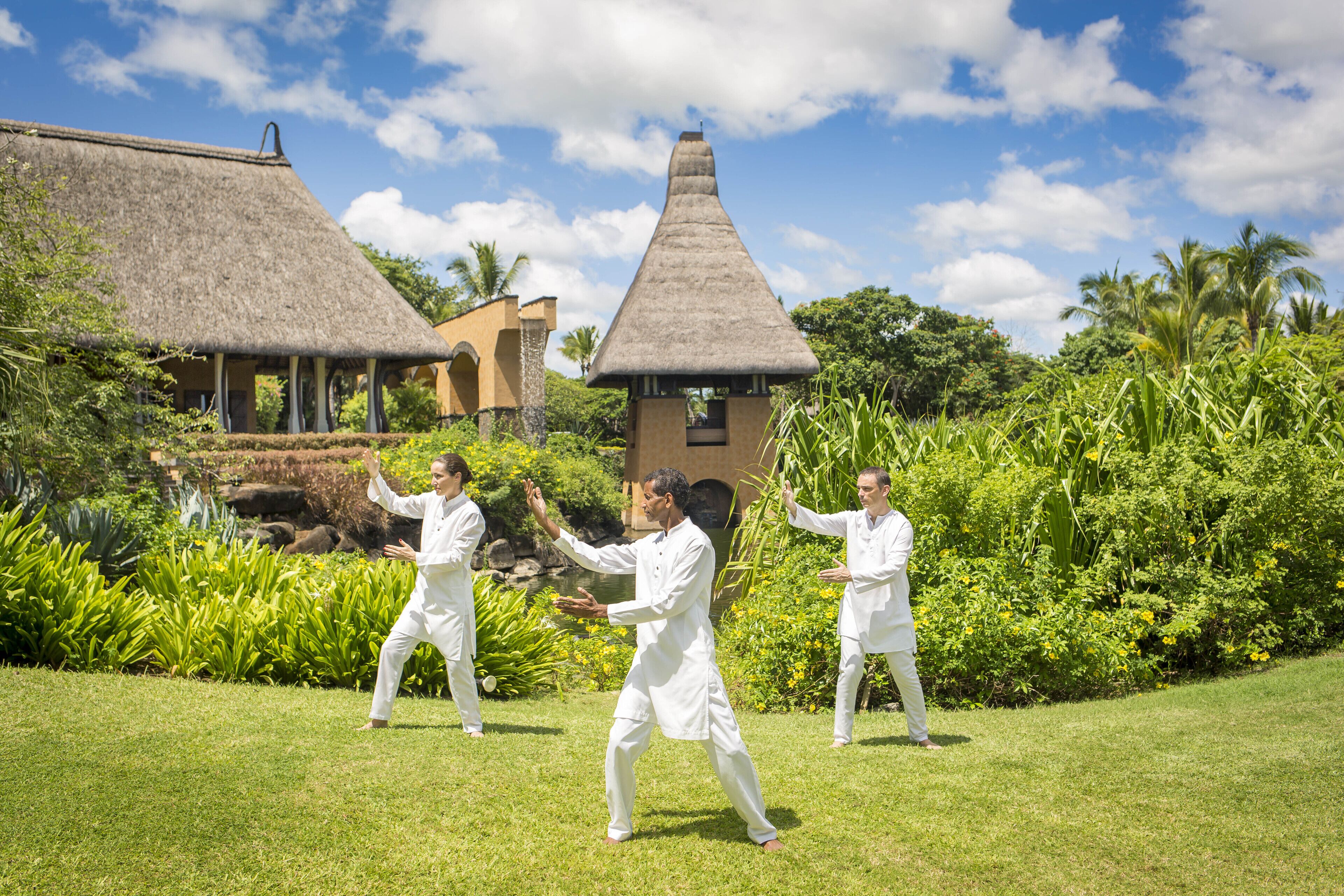 Un grupo de personas practicando artes marciales en un jardín exuberante, con una construcción de techo de paja al fondo.