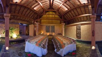 A room with a high, curved wooden ceiling and a central dome structure. There are two beds with floral bedspreads, a small water feature, and a wall with decorative elements.