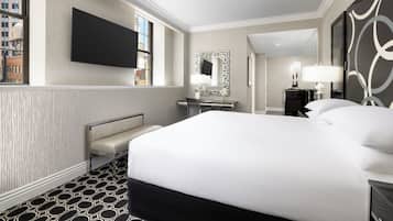 A hotel room with a large bed, a desk, a chair, a TV mounted on the wall, and a window with a view of buildings.