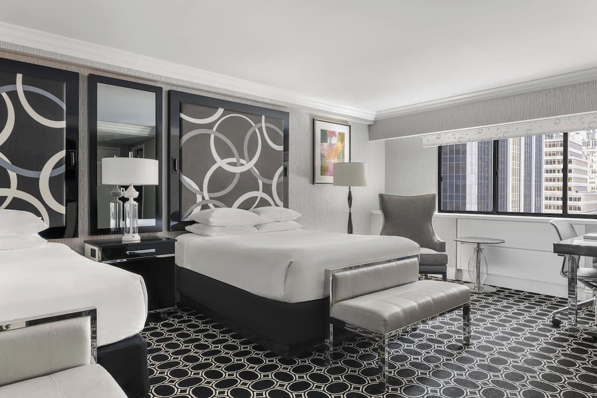 A modern hotel room with a large bed, a desk, a chair, and a view of the city through the window.