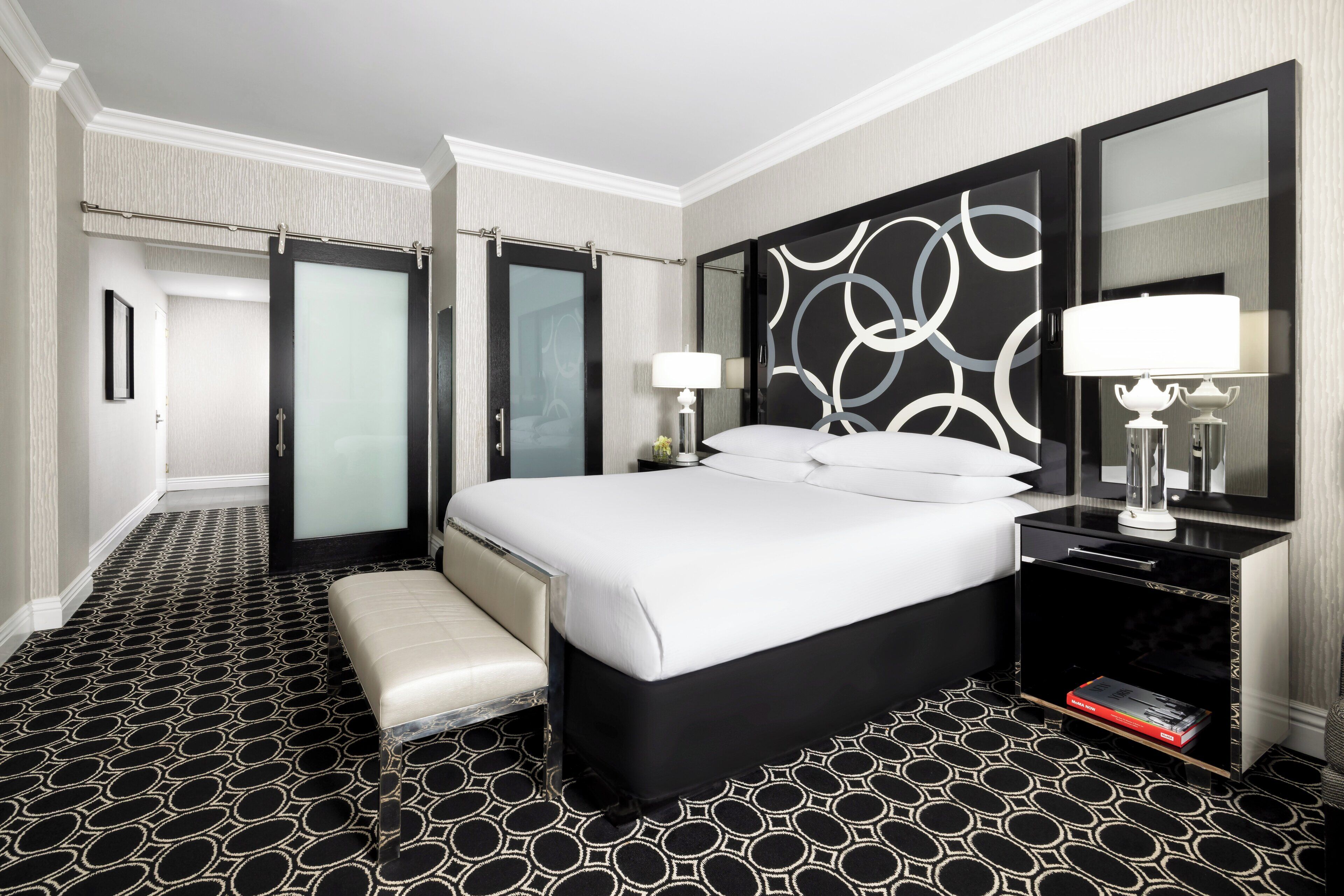A hotel room with a large bed, a bench, a bedside table with a lamp, and a mirror.