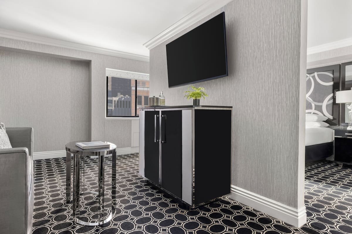 A hotel room with a large flat-screen TV mounted on the wall, a minibar, a small round table with a chair, and a patterned floor.