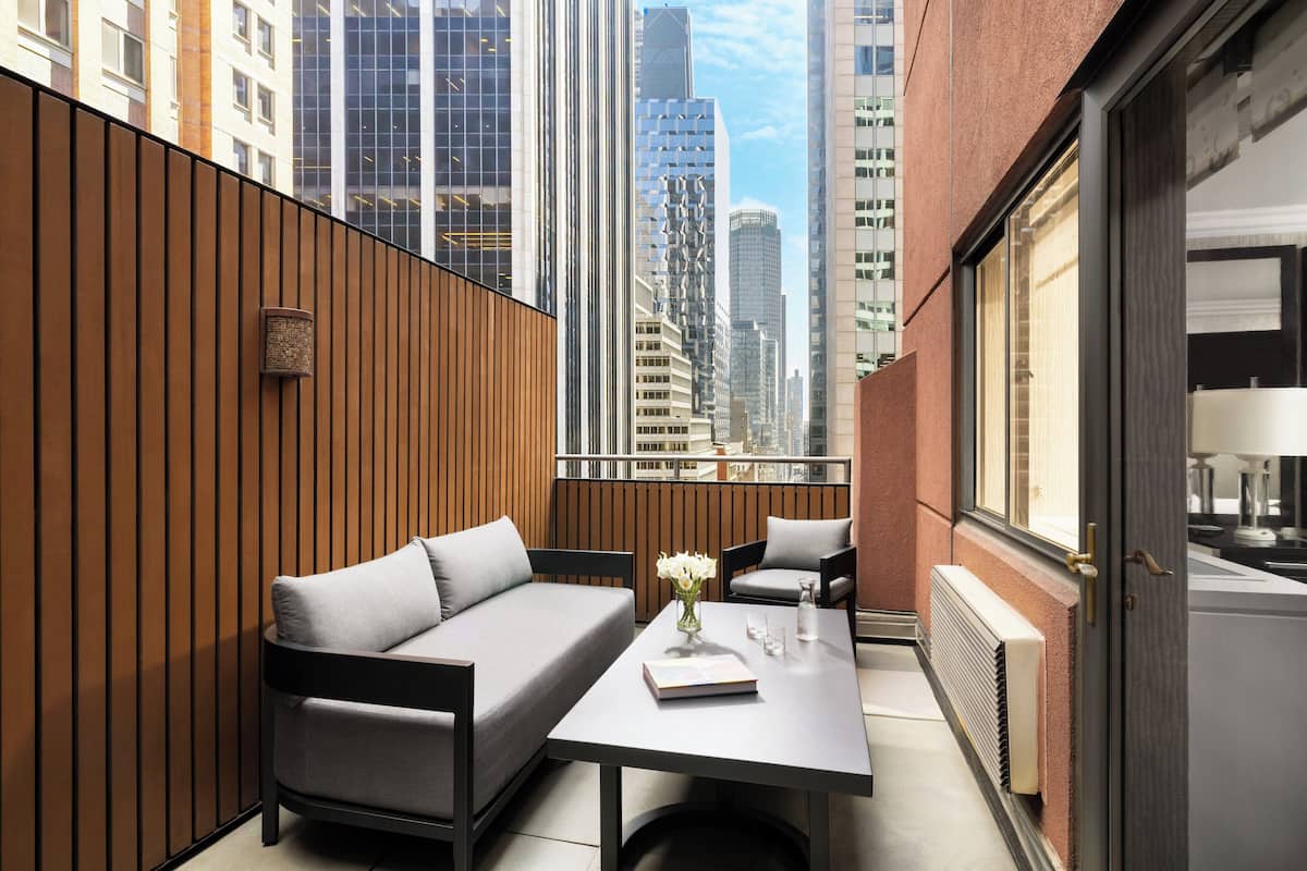 A modern urban balcony with a sofa, table, and chairs, overlooking a cityscape.