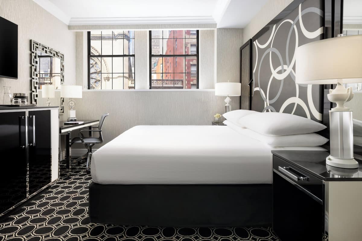 A hotel room with a large bed, a desk, a chair, a TV, and a window with a city view.